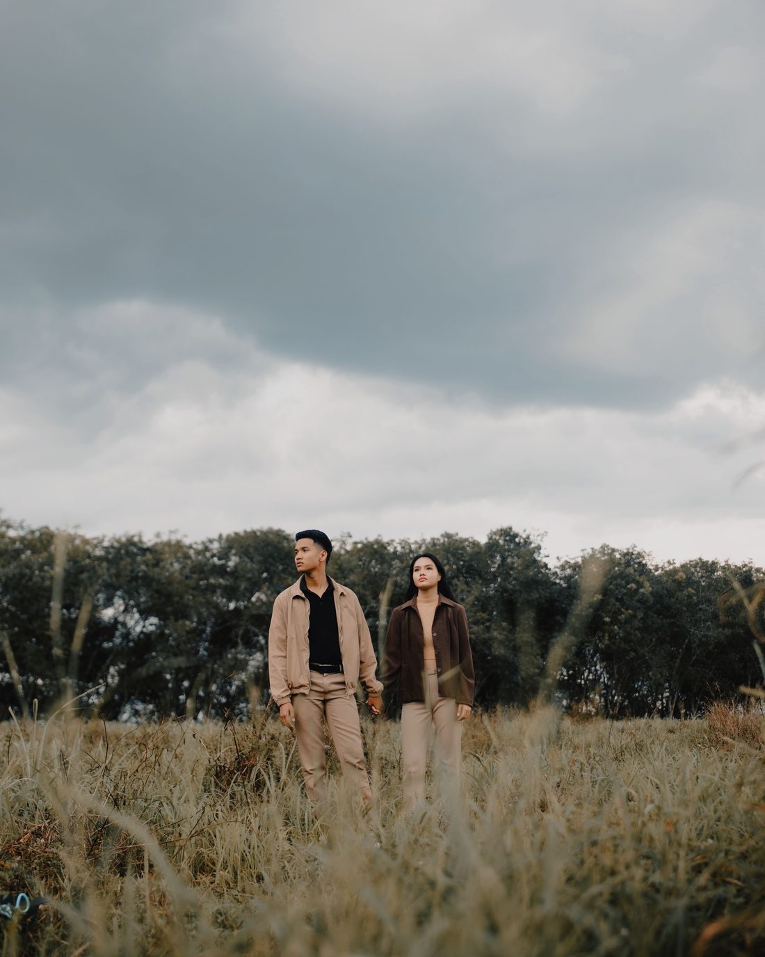 Twee mensen staan hand in hand in hoog gras onder een dramatische lucht